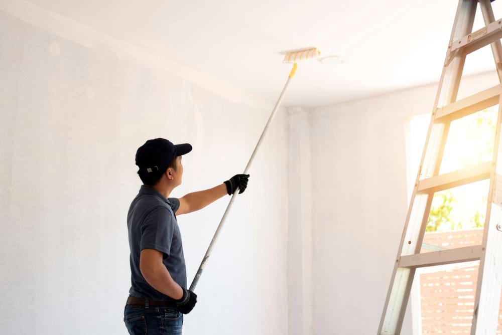 Asian rear view of a male painter drawing a wall with paint roller and a separate tank from a large empty space with wooden stairs