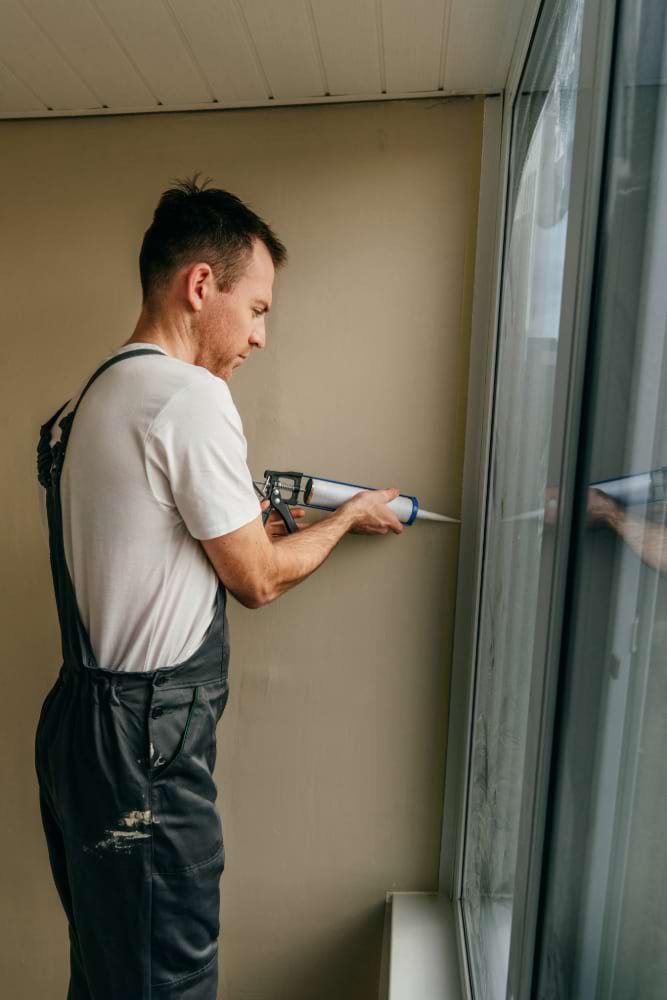Young man wearing overalls sealing cracks between window and trim