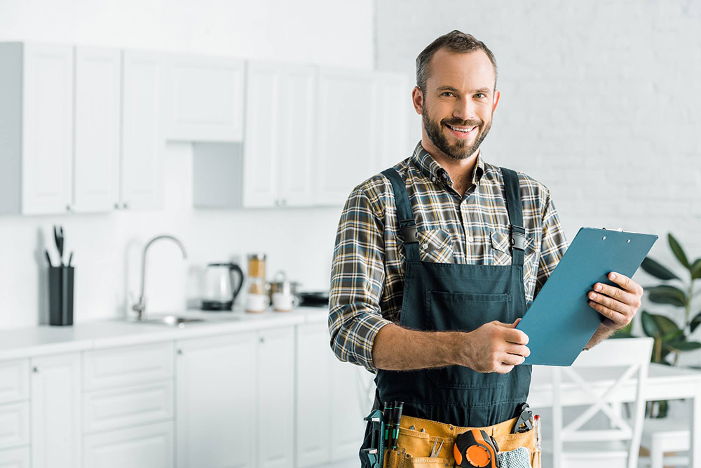 worker in overalls holding clipboard in white kitchen while facing camera and smiling