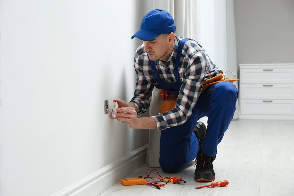 white male in blue overalls putting a new outlet fixture onto a white wall