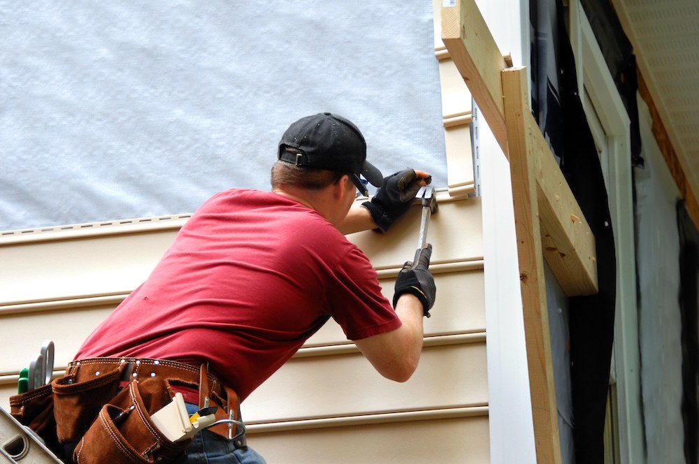 worker in red shirt and black baseball hat hammering on a roof